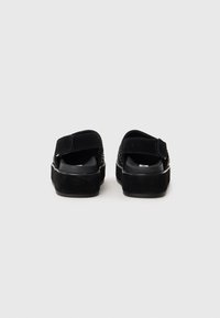 Schwarze Wildleder-Sandalen mit flacher Sohle, verstellbarem Klettverschluss und strukturierten Details an der Oberseite. Einfaches, modernes Design.