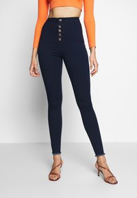 Jeans skinny de talle alto en azul marino con cierre de botones en la parte frontal y un dobladillo deshilachado sutil, combinados con una blusa corta de manga larga naranja.