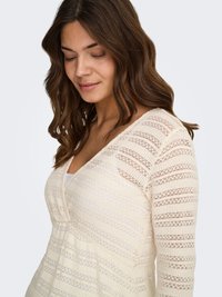 Ženska z valovitimi rjavimi lasmi, oblečena v kremno obarvan pleten top z nežnim črtastim vzorcem in v-izrezom, gleda navzdol z rahlim nasmeškom.