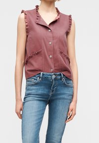 Haut sans manches mauve à boutons avec bords volantés, porté avec un jean denim bleu clair sur une personne debout.