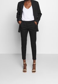 Kvinna klädd i svart blazer och byxor med vit V-ringad skjorta och beige högklackade sandaler med fotledsrem, med händerna i fickorna, stående mot vit bakgrund.