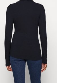 Svart ribbad långärmad topp med hög krage, med en figurnära passform och en lätt rundad nederkant. Kombinerad med mörk denimjeans.