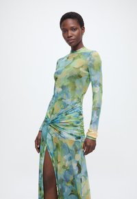 Γυναίκα που φοράει μακρυμάνικο φόρεμα με λουλουδάτο σχέδιο, με ραφές που δημιουργούν πιέτες και σκίσιμο μέχρι τον μηρό, ποζάροντας μπροστά σε λευκό, απλό φόντο.