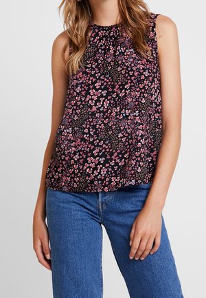 Bezrekawnikowa czarna bluzka z czerwono-różowym kwiatowym wzorem, noszona z niebieskimi dżinsami przez stojącą kobietę.