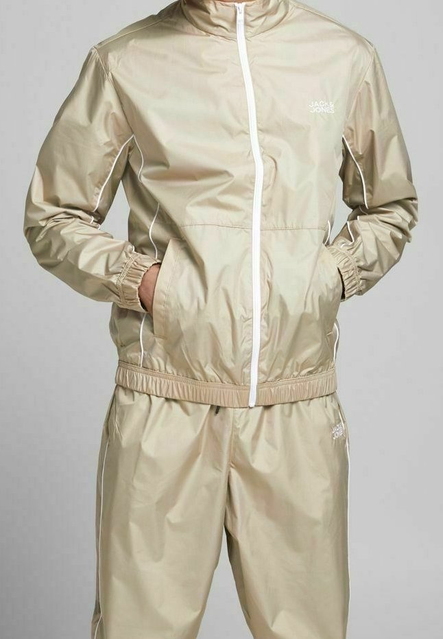 Beige vindjacka med vita detaljer, med hög krage, dragkedjestängning och elastiska ärmslut. Kombinerad med matchande byxor.