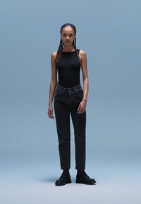 Zwarte bodysuit, hooggepollijste donkere jeans en zwarte legerboots. Het model heeft lang, gevlochten haar en staat tegen een lichtblauwe achtergrond.