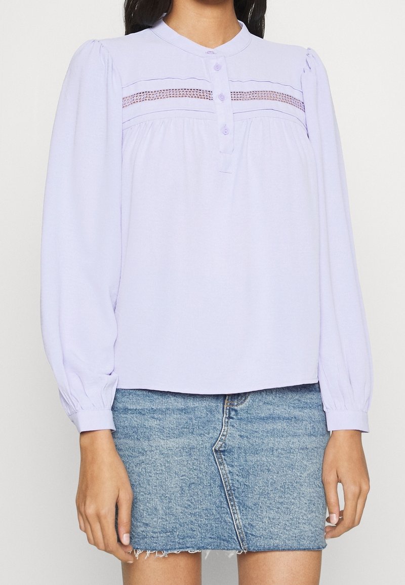 Blusa di un leggero viola con bottoni sul davanti, maniche a sbuffo e dettagli di smocking decorativi sul petto; abbinata a una gonna di denim.