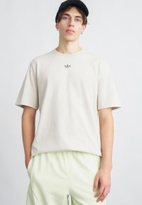 Tricou din bumbac bej cu mâneci scurte, având un mic logo gri pe piept. Purtați-l cu pantaloni cu talie elastică de culoare verde deschis.