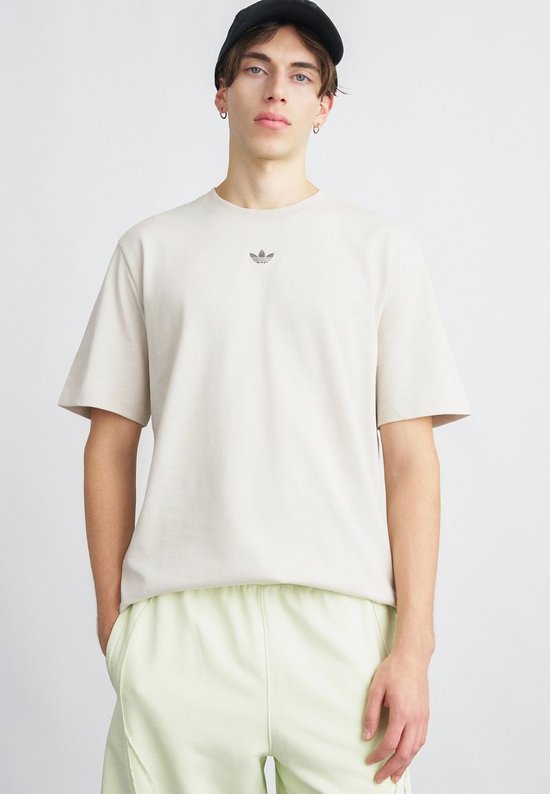 Tricou din bumbac bej cu mâneci scurte, având un mic logo gri pe piept. Purtați-l cu pantaloni cu talie elastică de culoare verde deschis.