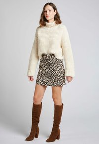 Pull en maille épaisse crémeux avec un col montant, associé à une mini jupe en imprimé léopard beige et des bottes en suède marron jusqu'au genou.