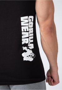Black sleeveless shirt made of cotton, featuring white text "GORILLA WEAR" and a gorilla graphic on the side. Simple design, casual fit.
