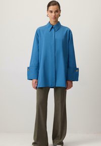 Sinine üle-suurusega nööpidega pluus pikemate varrukatega ja kuldsete nööbiaugukestega, kombineeritud kõrge vöökohaga oliivroheliste laiade pükstega.