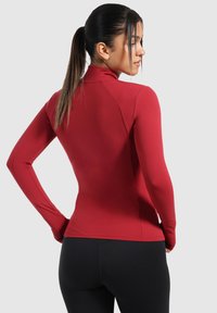Rød langærmet atletisk top med høj krave og tætsiddende design, lavet af glat, strækbart stof, parret med sorte leggings.