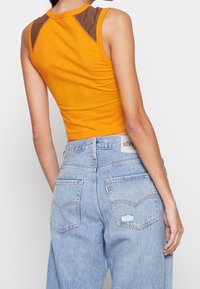 Gul kortärmad tanktopp med bruna axelaccenter, matchad med ljusblå denimhögmidjade jeans som har två bakfickor.