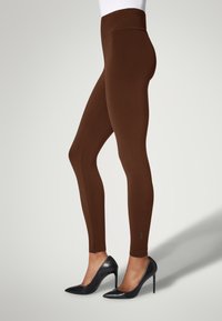 Bruna leggings med en slät textur, hög midja och en logodetalj vid ankeln, i kombination med glansiga svarta stilettklackar.