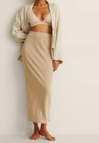 Beige ribbad lång kjol tillsammans med en matchande ljusbeige bralette och en lös beige kavaj, barfota mot en neutral bakgrund.