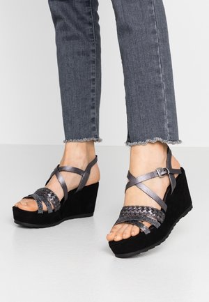 Pieds dans des sandales compensées noires avec des brides métalliques tressées et des boucles à la cheville, portées avec un jean gris foncé à ourlet effiloché.