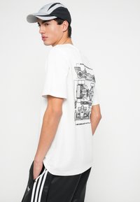 Baltas medvilninis marškinėlis su juodu grafiniu lenktyninių automobilių dizainu gale, derinamas su juodais sportiniais šortais ir pilka kepure.