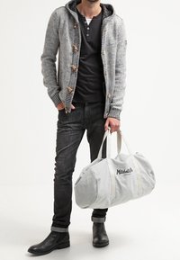 Cardigan grigio lavorato a maglia con cappuccio, bottoni in legno, camicia nera, jeans scuri, stivali neri e una borsa a tracolla grigia chiara con testo.