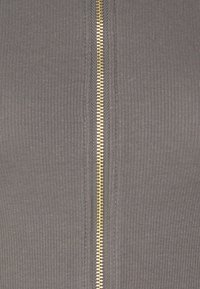 Vêtement zippé en tissu gris côtelé avec une fermeture éclair dorée centrale et des détails texturés sur les côtés.