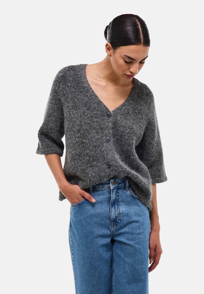 Pull en tricot gris oversize avec un décolleté en V profond, des manches trois-quarts et trois boutons de fermeture. Associé à un jean en denim bleu taille haute.