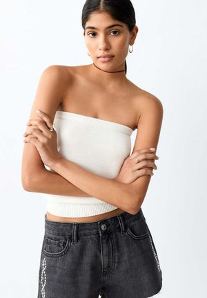 Femme aux cheveux foncés portant un top blanc sans bretelles et un jean noir, debout, les bras croisés, sur un fond blanc uni.