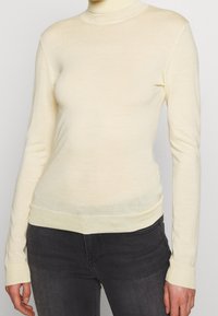 Femme portant un pull col roulé crème ajusté et un jean gris foncé devant un fond clair uni.