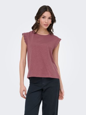 Woman wearing a mauve sleeveless top with lace-trimmed shoulders and black pants, standing against a plain light background.