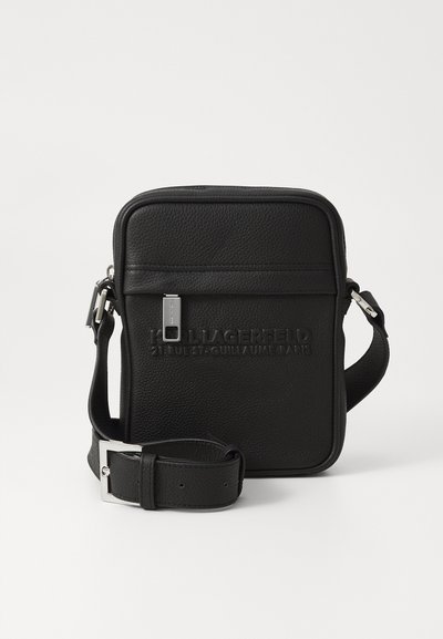 Sac en cuir noir à bandoulière avec logo embossé, quincaillerie en métal argenté, forme rectangulaire, sangle ajustable et surface texturée.