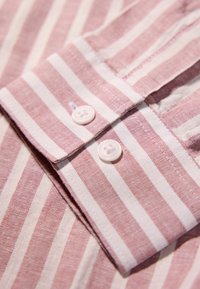 Camicia a righe con colori rosa chiaro e bianco, dal tessuto morbido. Colletto arrotondato con due bottoni e dettaglio di cucitura ben visibile.