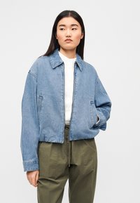 Μαλακό γαλάζιο τζιν μπουφάν με γιακά, εμπρόσθιο φερμουάρ και δύο πλαϊνές τσέπες, συνδυασμένο με λευκό πουκάμισο και παντελόνι χακί.