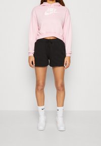 Růžová zkrácená mikina s bílým logem Nike, v kombinaci s černými šortkami se šňůrkou. Oblečení doplňují bílé ponožky a tenisky.