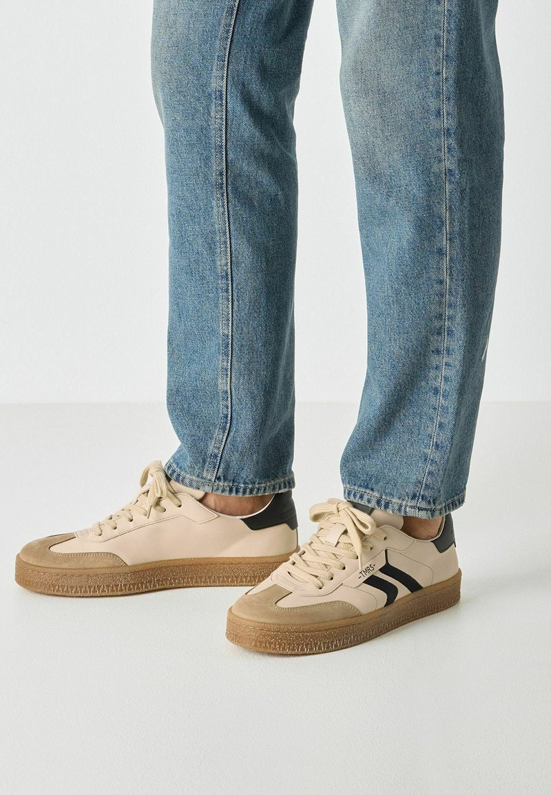 Beige sneakers with a smooth leather upper, black accents on the heel, and a textured brown rubber sole. Laces are cream-colored.