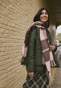 Green quilted jacket with a large brown and pink plaid scarf featuring fringe. The model wears a checkered long skirt with a slight shine.