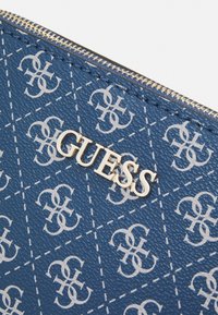 Blauwe gestructureerde portemonnee met een herhalend wit patroon en een gouden "GUESS" logo. Voorzien van een ritsluiting en gestikte accenten.