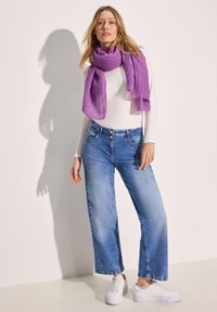 Sciarpa a coste viola, maglietta bianca a maniche lunghe, jeans a gamba larga azzurri chiari e sneakers bianche. Il modello è in piedi contro uno sfondo semplice.