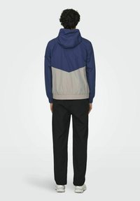 Windbreakerjack in marineblauw en lichtgrijs met een chevrondesign; heeft een capuchon, elastische manchetten en een gladde textuur. Zwarte broek en sneakers.