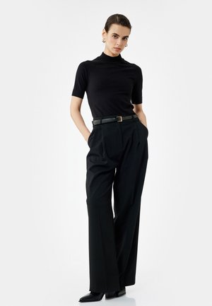 Schwarzes kurzärmliges Rollkragenoberteil kombiniert mit hoch taillierten schwarzen plissierten Hosen und einem schwarzen Gürtel mit einer goldenen Schnalle. Schwarze spitze Absatzschuhe.