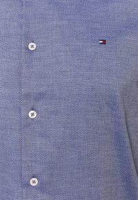 Camicetta di tessuto azzurro chiaro con bottoni bianchi e un piccolo logo ricamato rosso, bianco e blu sul petto.