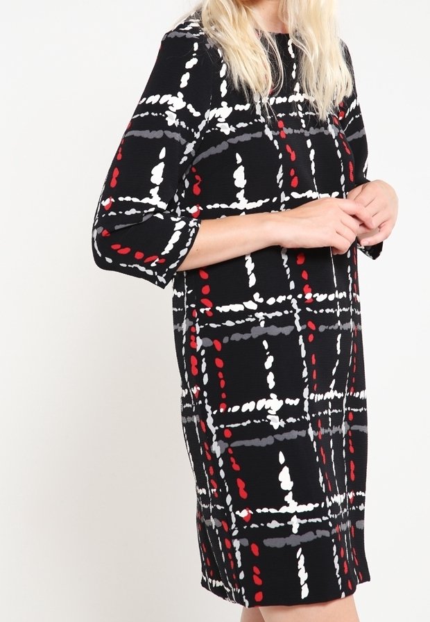 Femme portant une robe noire arrivant aux genoux avec un motif à carreaux abstrait blanc, rouge et gris et des manches trois-quarts.