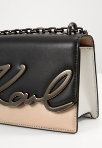 Bolso de mano de imitación de cuero negro y beige con un logo de firma en bronce, superficie texturizada, correa de cadena y forma estructurada.