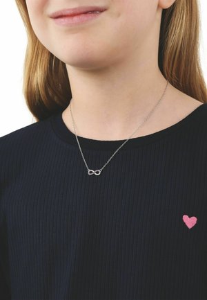 Giovane che indossa una collana con pendente a forma di infinito in argento e una maglia nera a coste con un piccolo ricamo a forma di cuore rosa.