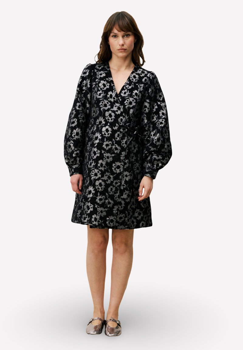 Zwarte jurk met bloemenpatroon, diepe V-hals, volumineuze mouwen en een getailleerde taille. De stof heeft een textuur. Zilverkleurige accenten op de schoenen.