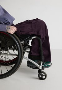 Pantalonii din catifea purpurie se așează peste un scaun cu rotile, având pliuri. Persoana poartă adidași verzi și negri. Detalii de aproape ale componentelor scaunului cu rotile sunt vizibile.