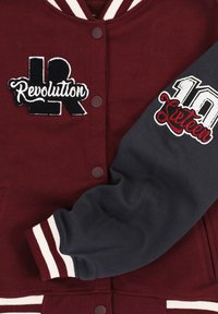 Bordeaux varsityjack met grijze mouwen, voorzien van een gestructureerde zwart-witte patch met de tekst "Revolution" en een rood-witte patch met "16".