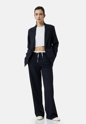 Mujer que lleva un blazer negro y pantalones de pierna ancha, top corto blanco, zapatillas blancas, de pie con las manos en los bolsillos contra un fondo blanco.