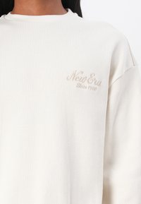 Cremfarbener, gerippter Sweatshirt mit Rundhalsausschnitt und gesticktem "New Era"-Logo in Beige auf der linken Brust.