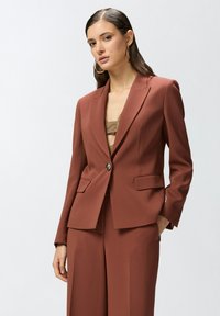 Smeđi sako prilagođenog kroja s jednim dugmetom, reverima i dva prednja džepa. Glatka tkanina s modernim fitom.