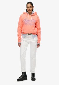 Koraliowy hoodie z nadrukowanym logo, białe spodnie i czarne sznurowane buty. Ubranie ma warstwową niebieską koszulę w paski pod hoodie.