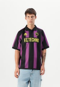 Mustapilkkuinen polo-paita, jossa on kaulus, ja jossa on keltaiset "F.C. Techno" -tekstit sekä pieni logo rinnassa. Siniset farkut näkyvät.